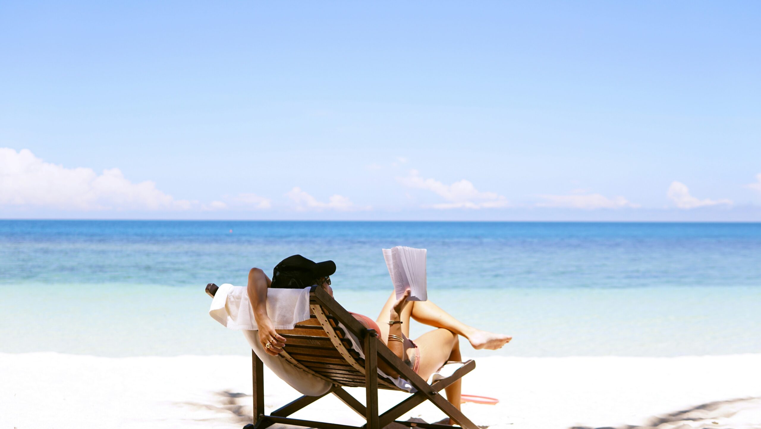 Person relaxing on a beach chair reading a book by the ocean, with a clear blue sky and calm sea in the background