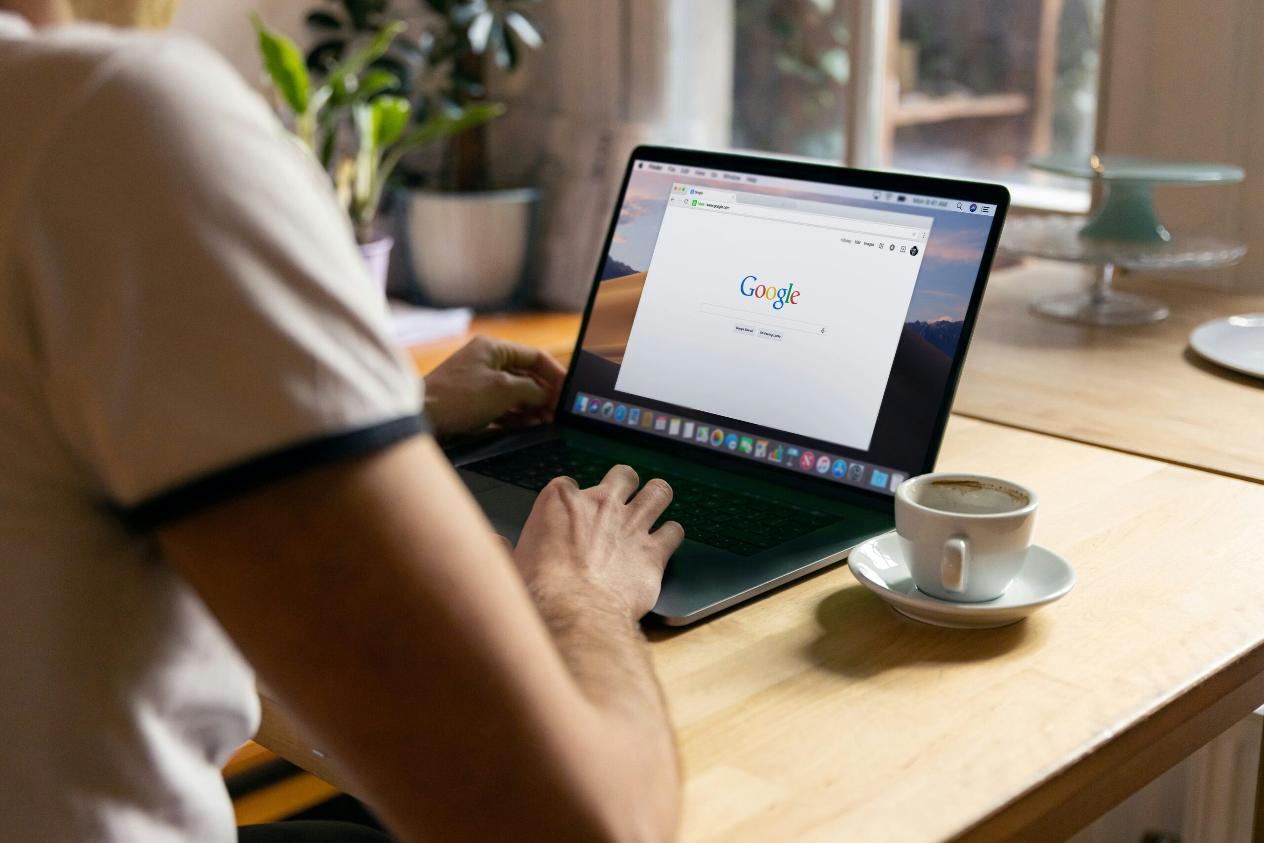 Person using a laptop with the Google search page open, sitting at a wooden desk with a cup of coffee nearby