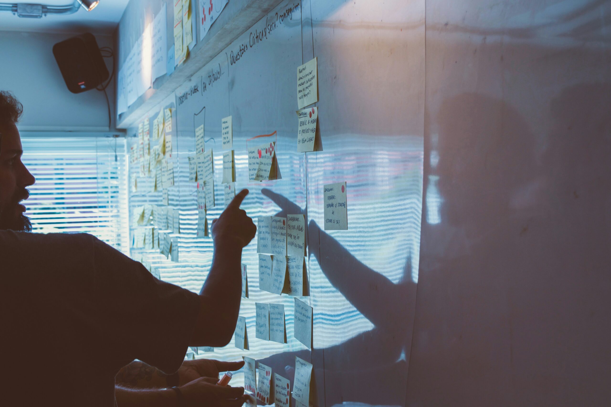 Person pointing at sticky notes on a whiteboard during a collaborative planning session, with light streaming through window blinds